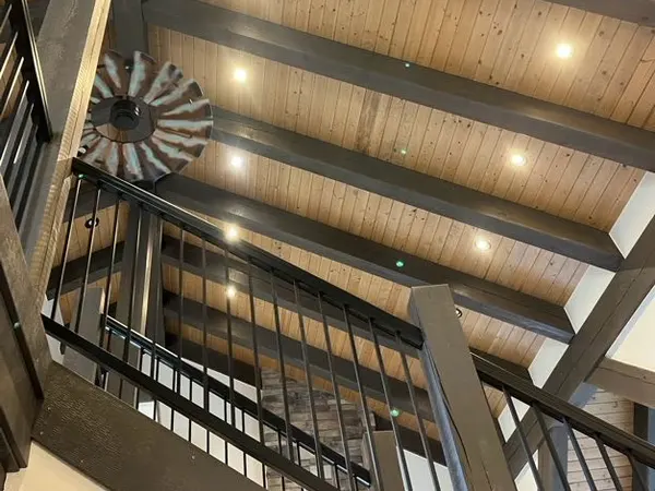 A stairwell under a plank ceiling lit by a row of recessed cans, with a windmill-blade fan on the beam above.