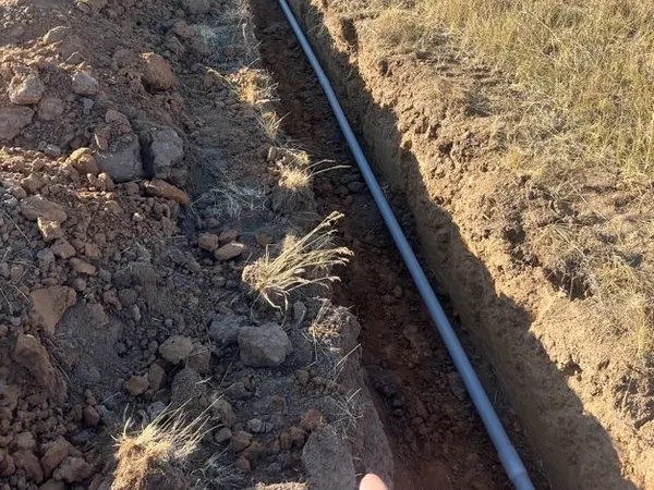 An open trench with gray conduit running across a field toward a distant house.