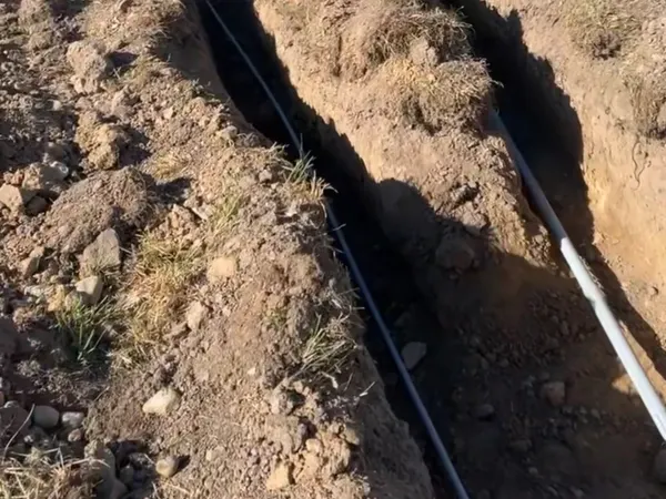 An open utility trench with buried conduit running across a rural field toward the Montana mountains.