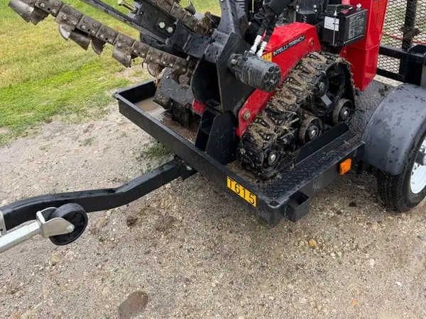 A walk-behind trencher on its trailer at a rural job site, staged beside a shop building and pines for an underground service run.
