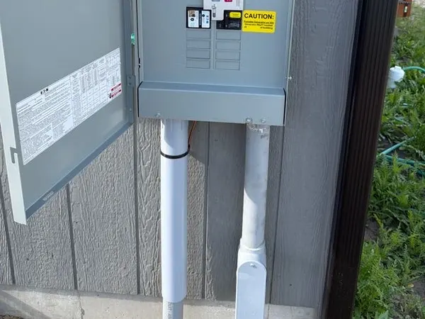 An open exterior home service panel with breakers and a conduit riser on lap siding.