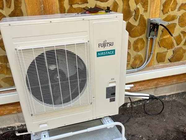 A Fujitsu mini-split condenser on a concrete pad with an electrical disconnect and conduit whip on a cabin wall.