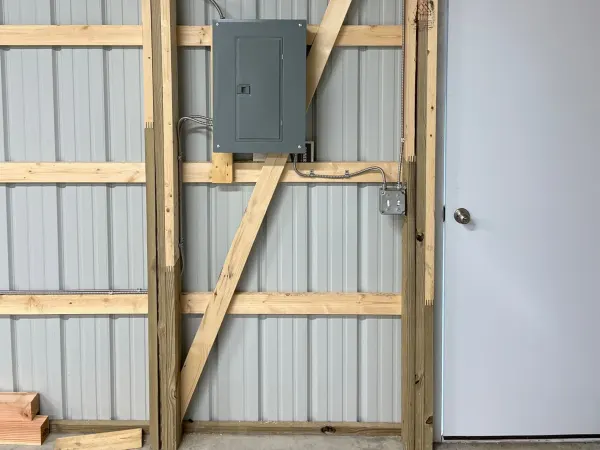 A gray breaker panel surface-mounted on wood framing in a metal pole-building, fed by armored cable, beside a service door.