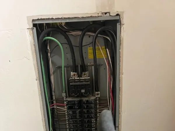A gloved electrician gives a thumbs-up in front of an open, neatly wired residential breaker panel.