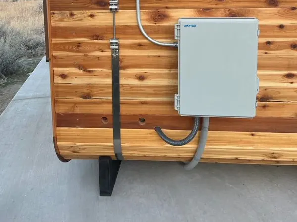 A surface-mounted electrical panel and conduit whip powering a cedar outdoor sauna, with the Bitterroot foothills behind.