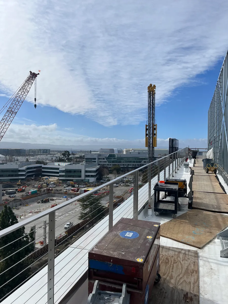 View from an upper floor of a large commercial building under construction, with a crawler crane, hoist tower, and materials staging yard below.